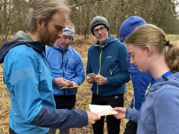 A group of orienteers are huddled, with the foreground showing a coach showing a map to a younger orienteer, providing coaching.